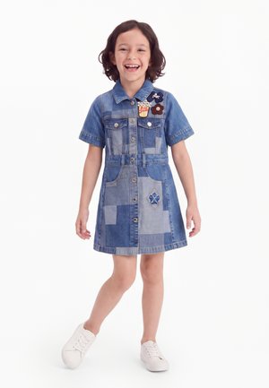 Lächelndes Mädchen steht mit einem Fuß nach vorne, trägt ein Patchwork-Denimkleid mit Blumen- und Popcornapplikationen und weiße Turnschuhe.