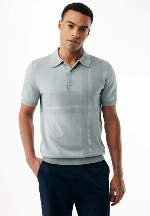 Giovane uomo con capelli corti e ricci che indossa una polo grigia chiara dalla trama strutturata e pantaloni scuri, in piedi con una mano in tasca.