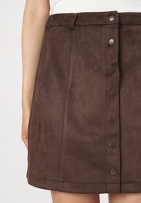 Närbild av en brun mocka mini kjol med en rad runda knappar framtill och bältesöglor, som bärs med en vit topp.