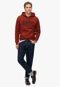 Hombre con sudadera de óxido con texto negro, jeans oscuros remangados, calcetines grises y zapatos negros, de pie con una mano en el bolsillo.