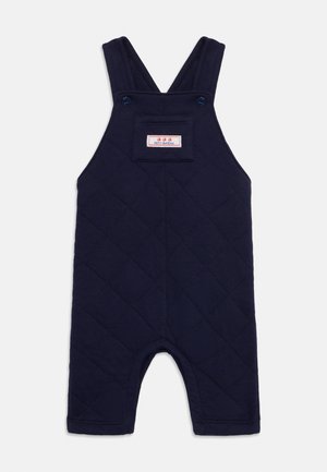 Navy blue quilted overall, featuring adjustable straps, a front pocket with a label, and hemmed edges. Cut-off legs for ease of movement.