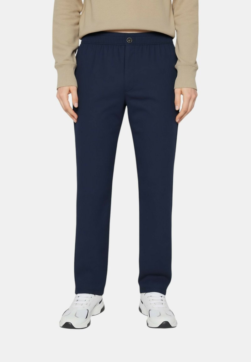 Pantaloni tuta blu navy con vita elastica e tasche. Il tessuto è morbido e ha una texture liscia. Indossati con scarpe da ginnastica bianche.