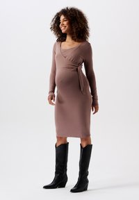 Robe de maternité en tissu mauve doux, avec un design enroulé, des manches longues et un lien à la taille, portée avec des bottes noires jusqu'au genou.