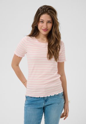 Jonge vrouw met lang golvend haar, gekleed in een roze-wit gestreept shirt met korte mouwen en een blauwe spijkerbroek, glimlachend met hand op heup.