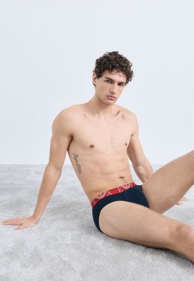 Cuecas azuis marinho com uma faixa elástica vermelha na cintura, com detalhe do logotipo. O tecido parece suave e ajustado, realçando a forma do corpo.