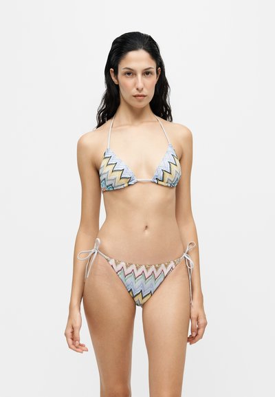 Femme aux longs cheveux foncés portant un bikini string à motif zigzag multicolore sur un fond clair uni.