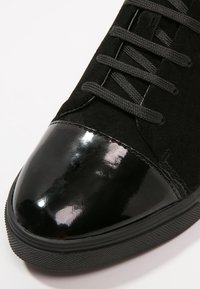 Černé tenisky s lesklým lakovaným špičkou a matným textilním svrškem, s černými tkaničkami a texturovanou gumovou podrážkou.