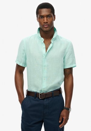 Camisa de lino de manga corta color azul claro con cuello mao y botones en la parte frontal, combinada con pantalones azul oscuro y un cinturón marrón.