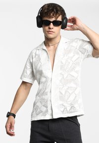 Witte shirt met korte mouwen en een geometrisch kantpatroon, met een relaxte pasvorm en een diepe V-hals. Gedragen met zwarte zonnebril en koptelefoon.