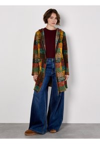 Kolorowy płaszcz z patchworku z kapturem w ciepłych odcieniach czerwieni, zieleni i brązu, w połączeniu z dżinsami o szerokich nogawkach i brązowym obuwiem.