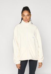 Kermanvärinen fleecetakki, jossa korkea kaulus, etuvetoketju ja joustavat hihansuut. Varustettu sivutaskulla ja kirjotulla logolla.