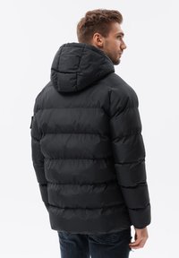 Schwarze, gepolsterte Jacke mit Kapuze, die ein gestepptes Design, glatte Textur und gerippte Bündchen aufweist. Die Rückansicht zeigt einen lässigen Schnitt und gestickte Details.