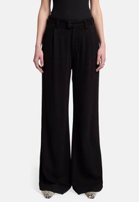 Pantalons larges noirs à taille haute, avec un devant plissé et un tissu lisse. Comprend des passants de ceinture et des poches latérales, se mariant bien avec des chaussures texturées.