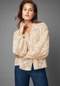 Cremefarvet bluse med blomstermønster, lange puffede ærmer, diskret knaplukning og en afslappet pasform, parret med mørk denimjeans.