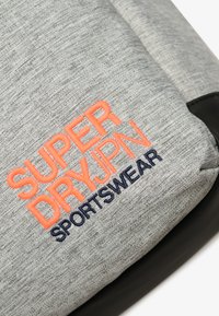 Tejido gris con el logo bordado "SUPER DRY JPN SPORTSWEAR" en naranja y negro sobre una superficie textil, mostrando detalles de textura y costura.
