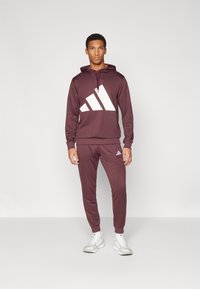 O hoodie atlético marrom apresenta um grande design gráfico branco. Combinado com calças de treino combinando. O tecido é suave, com uma silhueta ajustada.