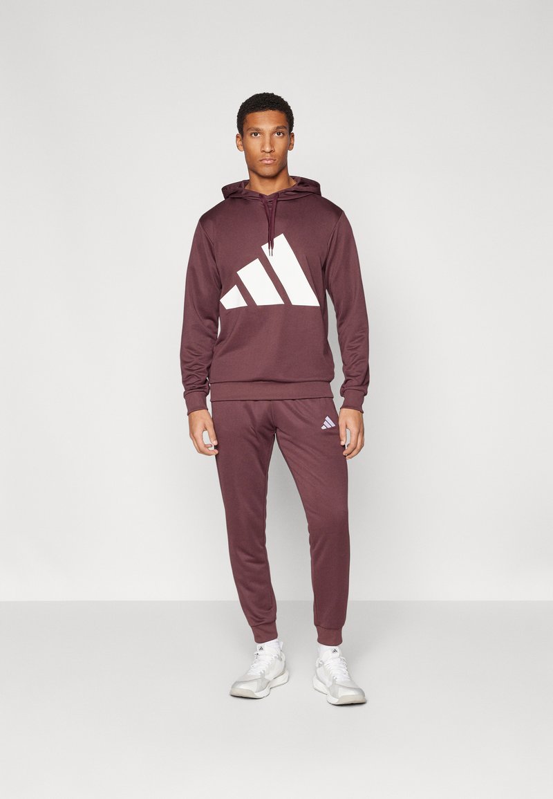 O hoodie atlético marrom apresenta um grande design gráfico branco. Combinado com calças de treino combinando. O tecido é suave, com uma silhueta ajustada.