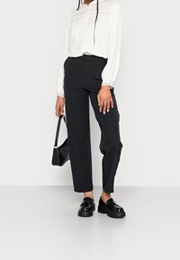 Krämfärgad blus med knappar, spetsdetalj, svarta högmidjade byxor och glänsande svarta loafers. Liten svart handväska med rem.