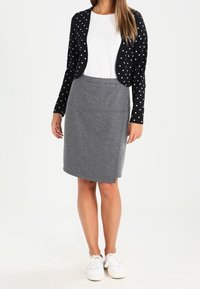 Cardigan nero con pois bianchi sopra una maglia bianca strutturata, abbinato a una gonna grigia fino al ginocchio e sneakers bianche. Tessuto liscio, stile casual.