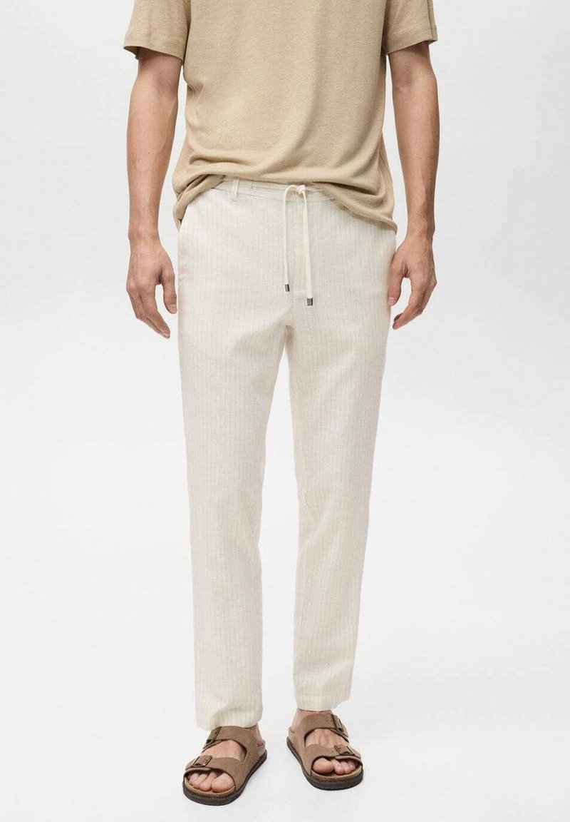 Homme portant une chemise beige à manches courtes, un pantalon à cordon blanc cassé et des sandales marron à bouts ouverts, sur un fond blanc uni.