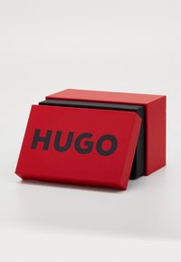 Boîte rectangulaire rouge avec un intérieur noir et un couvercle affichant le mot "HUGO" en lettres noires en gras. Texture matte lisse.