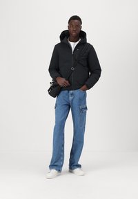 Doudoune noire avec capuche, jean cargo bleu à jambes larges et baskets blanches. Sac à bandoulière noir avec bandoulière ajustable.