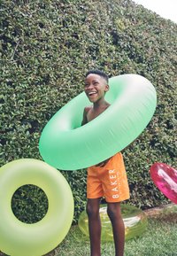 Dječak u narančastim kupaćim hlačicama smiješi se na otvorenom s velikim zelenim napuhanim prstenom za plivanje oko tijela, u blizini drugih šarenih prstenova za plivanje.