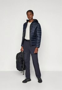 Marineblaue Steppjacke mit Kapuze, horizontal gestepptes Design, kombiniert mit dunkler Hose und schwarzem Rucksack, stehend vor neutralem Hintergrund.