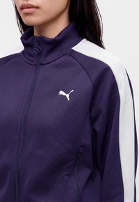 Γυναίκα με μωβ αθλητικό τζάκετ Puma με φερμουάρ και λευκές ρίγες στους ώμους· διακρίνεται μόνο το κάτω μέρος του προσώπου και το πάνω μέρος του σώματος.