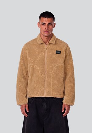 Mann trägt eine beige strukturierte Fleecejacke mit Reißverschluss und kleinem schwarzem Logo-Patch auf der Brust sowie schwarze Hose vor neutralem Hintergrund.