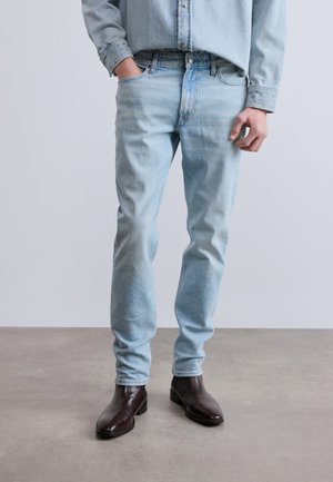 Personne portant un jean bleu clair, une chemise en denim assortie et des bottes en cuir marron foncé, debout sur un sol gris.