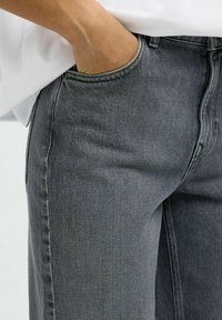 Mano derecha metida en el bolsillo delantero de unos vaqueros de mezclilla grises, llevados con una camisa blanca.