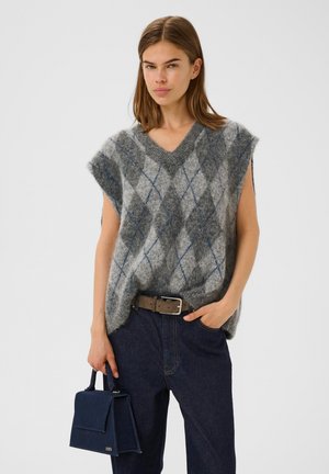 Grauer gemusterter oversized Strickwesten mit V-Ausschnitt, verziert mit blauen Diamantakzenten, getragen über dunklen Denim-Hosen, begleitet von einer kleinen marineblauen Handtasche.