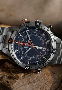 Montre Timex Expedition avec un cadran bleu marine, un bracelet en acier inoxydable argenté, des index horaires lumineux, et un indicateur de compas et de marées orange.