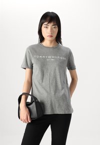 Szürke pamut póló rövid ujjakkal és kerek nyakkivágással, a fehér "TOMMY HILFIGER EST. 1985" felirattal, fekete kézitáskával párosítva.