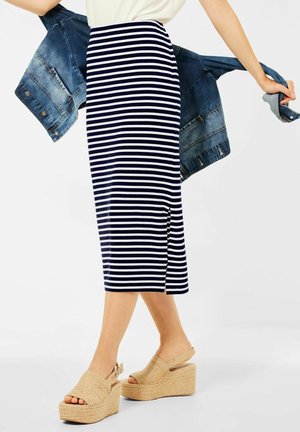 Femme portant une jupe midi rayée bleu marine et blanche ainsi que des sandales compensées beiges, tenant une veste en jean bleue derrière son dos.