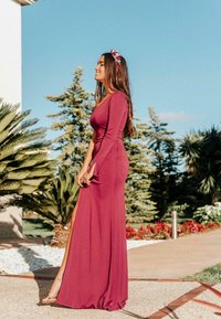 Largo vestido burdeos con un corsé ajustado y mangas largas, que presenta una gran abertura. La modelo lleva una corona de flores y se muestra al aire libre.