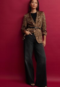Blazer à imprimé léopard en tissu lisse, associé à un jean noir à taille haute et jambes larges. La personne tient sa poche, se tenant devant un fond rose.