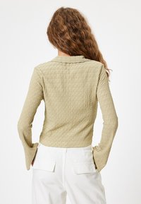 Haut à manches longues en vert olive clair avec un motif ondulé texturé. Présente un col et des poignets de manches évasés. Associé à un pantalon blanc.