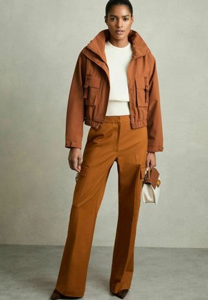 Giacca corta marrone con due tasche anteriori, maglione bianco sotto, abbinata a pantaloni cargo marroni a gamba larga e una borsa piccola.