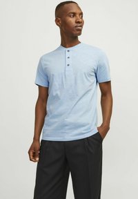 Camisa henley de manga corta azul claro con tres botones oscuros en el cuello, combinada con pantalones negros con pliegues. Textura suave.
