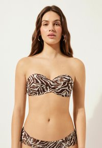 Avtagbar bikiniöverdel i brun och beige zebramönster med tvinnad detalj fram. Slät, elastisk tyg. Accentuerad med medelstora hoops örhängen.