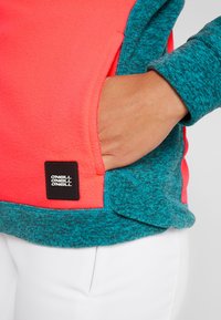 Sudadera de forro polar con un cuerpo de color rosa brillante y acentos en color teal. Cuenta con un bolsillo lateral y una etiqueta negra con texto. Textura suave en todo el producto.