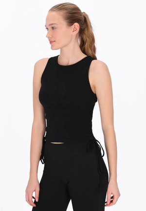 Fiatal nő világosbarna copffal, fekete, testhezálló ujjatlan felsőt és fekete nadrágot visel, balra nézve, fehér háttér előtt.