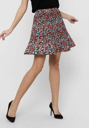 Femme portant une jupe fleurie multicolore mi-longue, un haut noir et des chaussures à talons noires, posant jambes croisées et bras étendus.