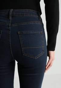 Tmavě modré džíny s vysokým pasem, s kontrastním šitím a dvěma zadními kapsami. Hladká textura a přiléhavý design.