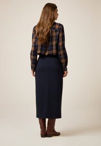 Karohemd in Marineblau und Braun, kombiniert mit einem langen Rock in dunklem Marineblau. Braune kniehohe Stiefel. Glatte Textur und taillierte Passform.