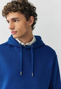 Blaues Kapuzensweatshirt aus weichem Stoff, mit Kordeln mit metallischen Enden und lockerer Passform, über einem weißen, mit Kragen versehenen Hemd getragen.