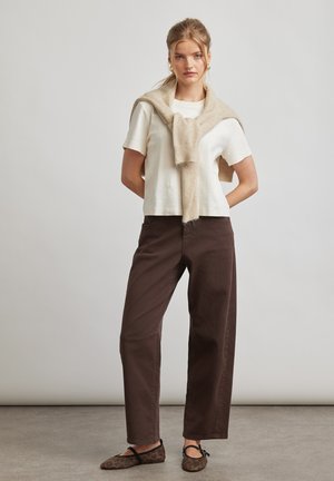 Frau steht mit den Händen hinter dem Rücken, trägt ein weißes Kurzarmshirt, einen beigen Pullover über den Schultern und dunkelbraune Hosen sowie gemusterte Ballerinas.