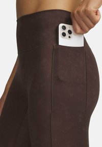 Phone with triple camera held in hand, partially inserted into the back pocket of dark brown fitted leggings.